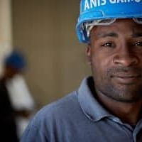 Portrait d’un homme portant un casque de chantier bleu, regardant l’objectif, avec un autre travailleur flou en arrière-plan.